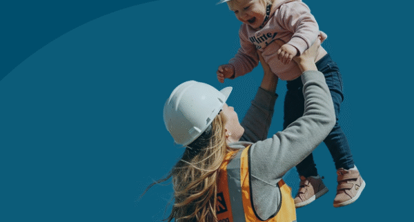 A woman wearing a high-visibility orange safety vest and a white construction helmet lifts a smiling toddler into the air. The toddler, also wearing a small white hard hat, pink jumper, and jeans, laughs joyfully against a plain blue background.