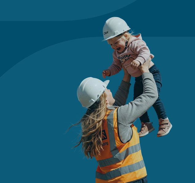 A woman wearing a high-visibility orange safety vest and a white construction helmet lifts a smiling toddler into the air. The toddler, also wearing a small white hard hat, pink jumper, and jeans, laughs joyfully against a plain blue background.