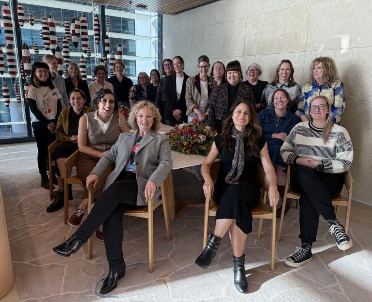Group photo of the Working Women’s Centre Network. About 21 people are gathered indoors, smiling at the camera. A large table with a flower arrangement sits in the middle, and red-and-white hanging decorations and city buildings are visible through the window in the background.