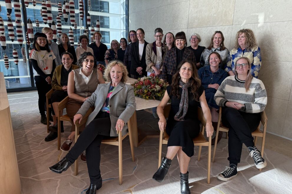 Group photo of the Working Women’s Centre Network. About 21 people are gathered indoors, smiling at the camera. A large table with a flower arrangement sits in the middle, and red-and-white hanging decorations and city buildings are visible through the window in the background.