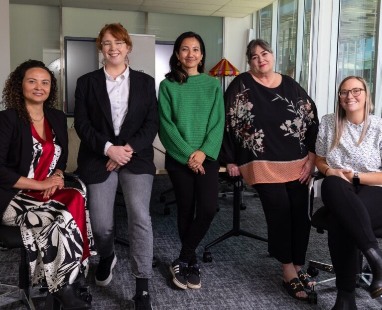 Five team members of the Working Women’s Centre Australia standing and sitting together in an office space, smiling at the camera.