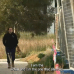 A woman wearing a dark jumper and work boots walks alone along a suburban footpath bordered by tall grass and construction materials. She appears thoughtful as she moves past a row of unfinished buildings. Subtitles on the image read, “I am the victim and I’m the one that’s silenced.”