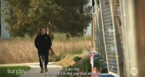 A woman wearing a dark jumper and work boots walks alone along a suburban footpath bordered by tall grass and construction materials. She appears thoughtful as she moves past a row of unfinished buildings. Subtitles on the image read, “I am the victim and I’m the one that’s silenced.”