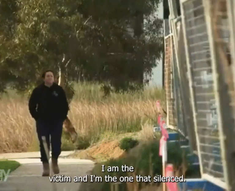 A woman wearing a dark jumper and work boots walks alone along a suburban footpath bordered by tall grass and construction materials. She appears thoughtful as she moves past a row of unfinished buildings. Subtitles on the image read, “I am the victim and I’m the one that’s silenced.”