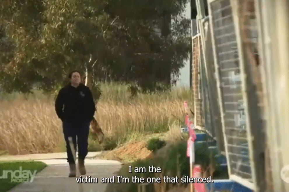 A woman wearing a dark jumper and work boots walks alone along a suburban footpath bordered by tall grass and construction materials. She appears thoughtful as she moves past a row of unfinished buildings. Subtitles on the image read, “I am the victim and I’m the one that’s silenced.”
