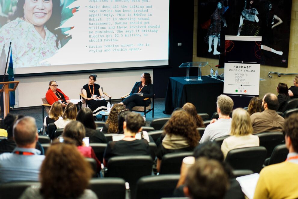 Panel of three women speak on stage about sexual harassment as audience listens in a lecture hall.