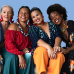 Four women of different ages and ethnic backgrounds sit closely together on a couch against a plain blue background, smiling warmly at the camera. They wear colourful, patterned clothing and statement earrings, leaning into one another in a relaxed, friendly pose that conveys connection and solidarity.