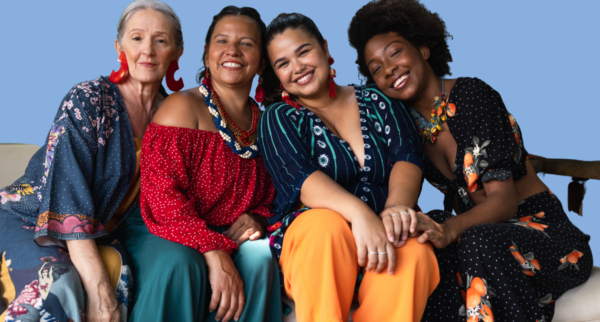 Four women of different ages and ethnic backgrounds sit closely together on a couch against a plain blue background, smiling warmly at the camera. They wear colourful, patterned clothing and statement earrings, leaning into one another in a relaxed, friendly pose that conveys connection and solidarity.