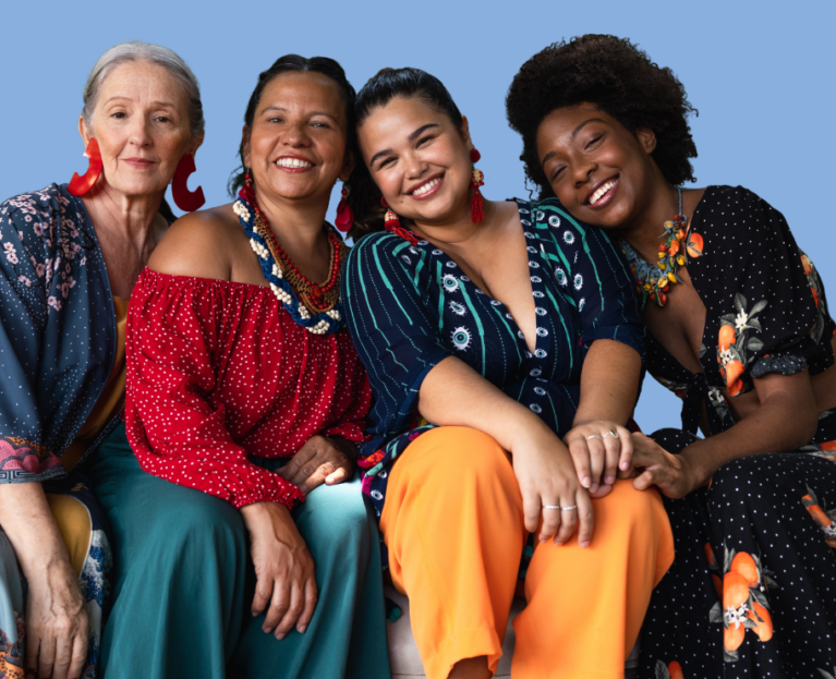 Four women of different ages and ethnic backgrounds sit closely together on a couch against a plain blue background, smiling warmly at the camera. They wear colourful, patterned clothing and statement earrings, leaning into one another in a relaxed, friendly pose that conveys connection and solidarity.