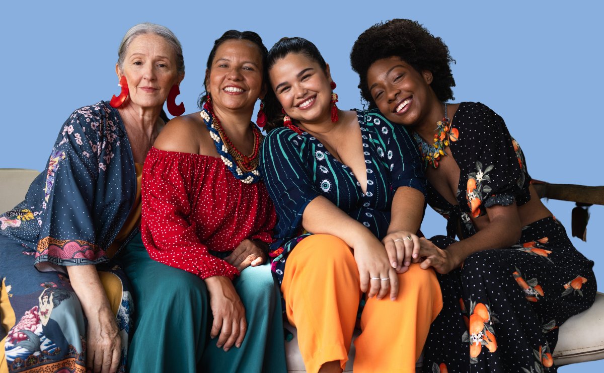 Four women of different ages and ethnic backgrounds sit closely together on a couch against a plain blue background, smiling warmly at the camera. They wear colourful, patterned clothing and statement earrings, leaning into one another in a relaxed, friendly pose that conveys connection and solidarity.