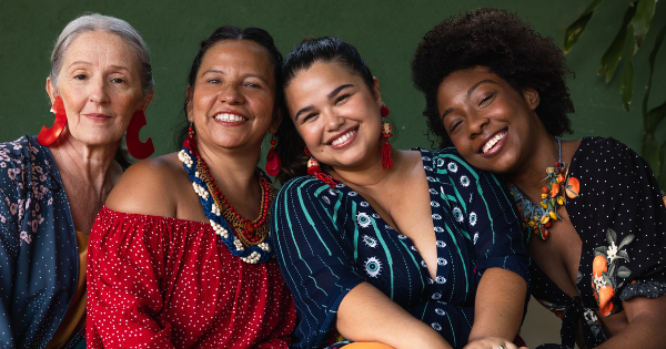 Four women of different ages and backgrounds sit closely together, smiling warmly at the camera, wearing colourful clothing and jewellery against a green background.