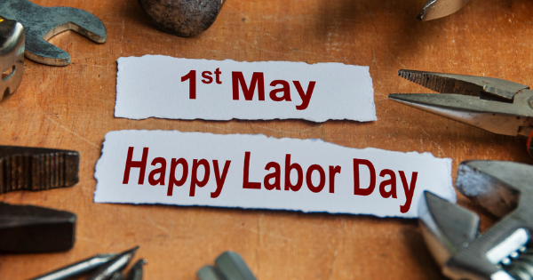 Close-up of a workbench with tools scattered around paper labels reading “1st May” and “Happy Labor Day.”